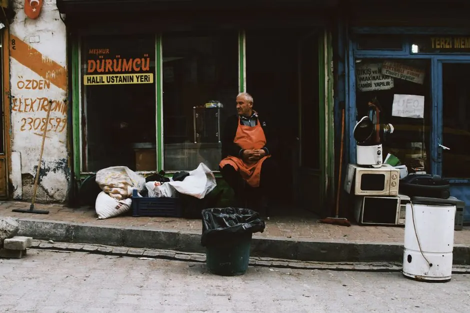 Shopkeeper in an apron sits outside a store in a bustling urban area.